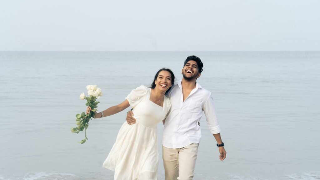 pexels-photo-30488053-30488053 Happy couple celebrating love by the sea, holding flowers and smiling.